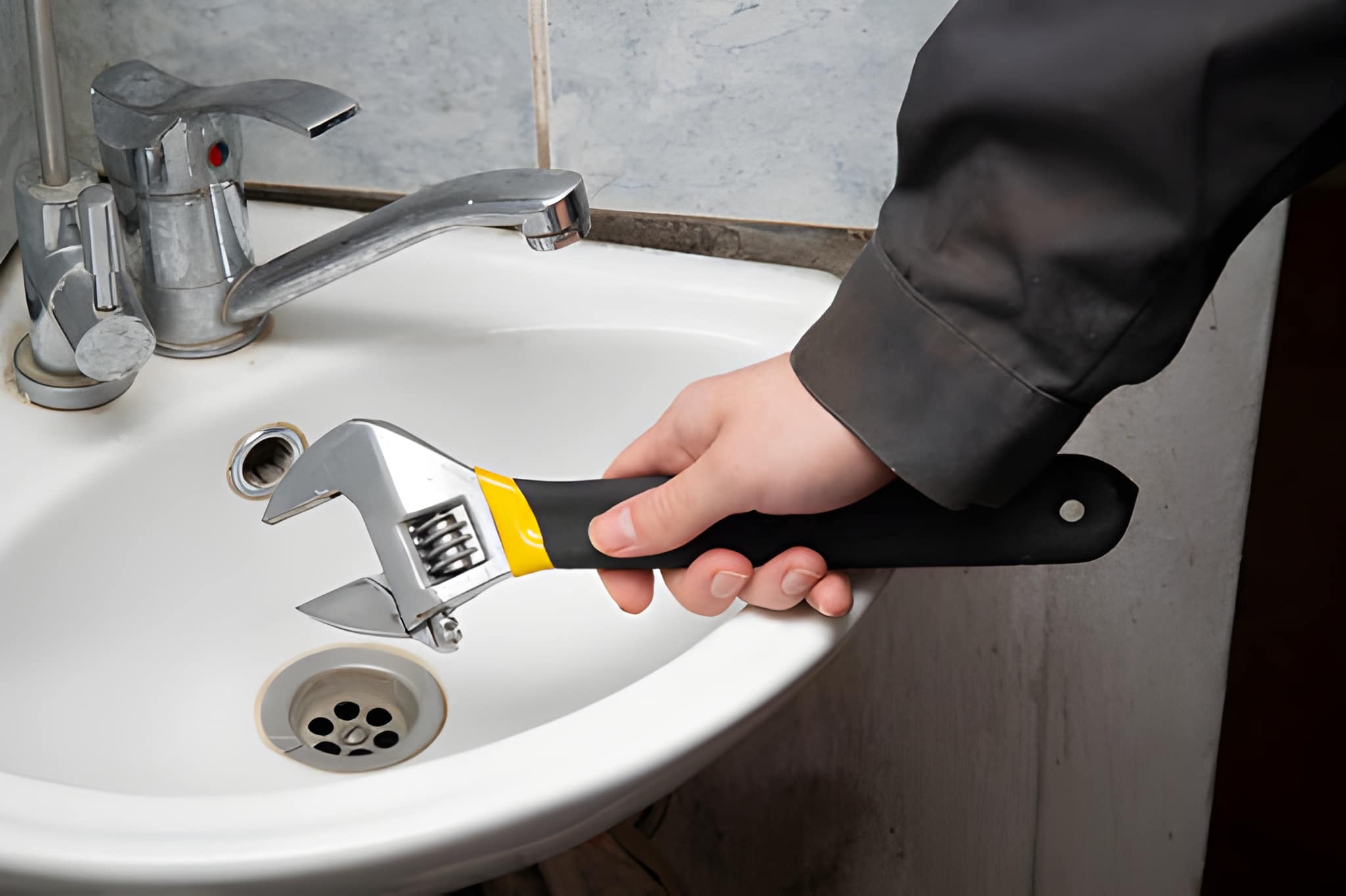 Mano sosteniendo una llave inglesa sobre un lavabo de baño blanco.
